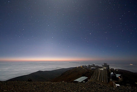 A dark blue starry sky. with light on the horizon, over a plain and an observatory.